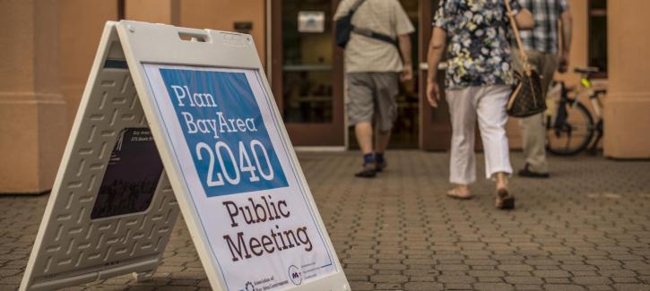 In the foreground a sandwich board advertising the Plan Bay Area 2040 public meeting is displayed, with three adults walking into the building in the background.